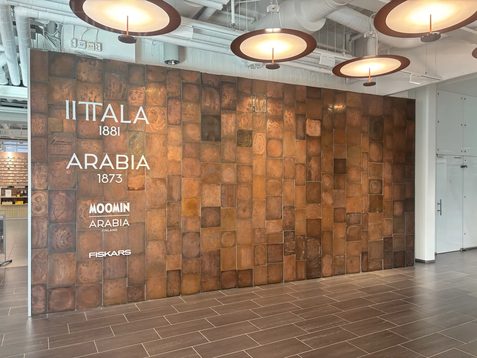 IITTALA 1881、ARABIA 1873、MOOMIN ARABIA、FISKARS のロゴが並ぶ銅板の壁